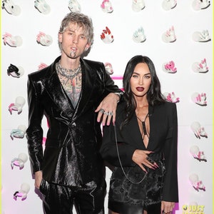 May include: A couple poses in front of a wall decorated with white hands. The man is wearing a black leather suit and has a silver chain necklace. The woman is wearing a black blazer over a black dress with a plunging neckline. Both have long, dark nails.