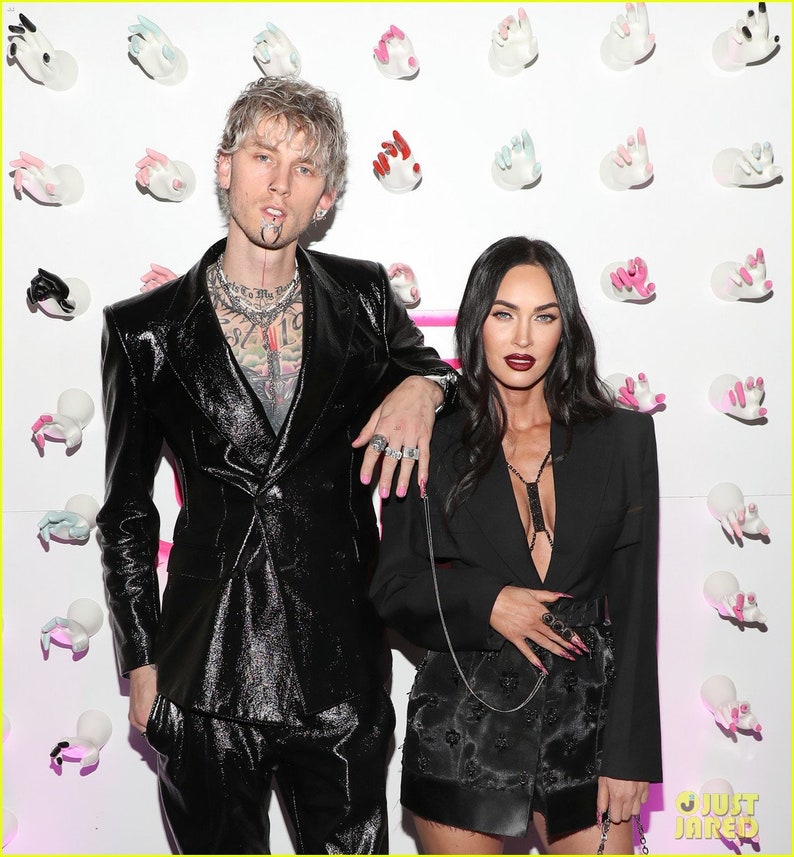 May include: A couple poses in front of a wall decorated with white hands. The man is wearing a black leather suit and has a silver chain necklace. The woman is wearing a black blazer over a black dress with a plunging neckline. Both have long, dark nails.