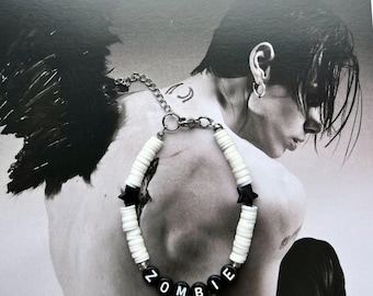 May include: A white and black beaded bracelet with the word "ZOMBIE" spelled out in black letter beads. The bracelet features black star-shaped beads and a silver chain clasp. The background is a black and white photograph.