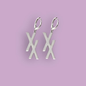 以下が含まれることがあります： シルバーカラーのイヤリングのペア。各イヤリングは、様式化された文字「X」のデザインが特徴です。イヤリングは、シルバーのレバーバッククロージャーから吊り下げられています。背景は無地のラベンダー色です。