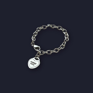 May include: Silver-toned bracelet with a chain link design. A heart-shaped charm dangles from the bracelet, with the words "GIMME KISS" inscribed in black. The bracelet is set against a dark background.