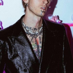 May include: A man wearing a black velvet blazer with multiple silver chains and a pearl necklace. He has a tattoo on his chest that says "Backs To My Youth" and other tattoos on his chest and neck.