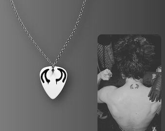 May include: A silver-toned necklace with a white pendant featuring a black, stylized design. The pendant hangs from a delicate chain. A black and white image of a person with a matching tattoo is in the background.