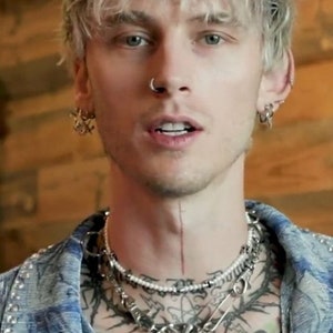 May include: A close-up portrait of a person with short, light blonde hair. They are wearing a blue denim jacket with rhinestones and have multiple silver necklaces with a large silver pendant. They have a nose ring and multiple earrings in both ears.