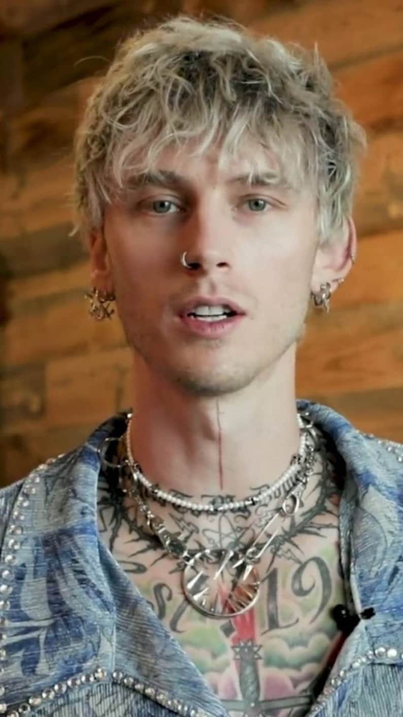 May include: A close-up portrait of a person with short, light blonde hair. They are wearing a blue denim jacket with rhinestones and have multiple silver necklaces with a large silver pendant. They have a nose ring and multiple earrings in both ears.