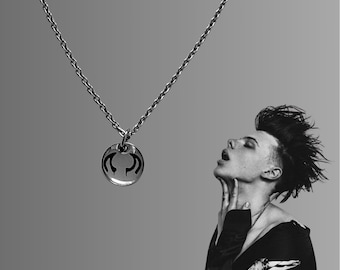 May include: Silver-toned necklace with a circular pendant featuring a stylized design. The chain is a classic link style. The image is a black and white portrait of a person with their head tilted back.