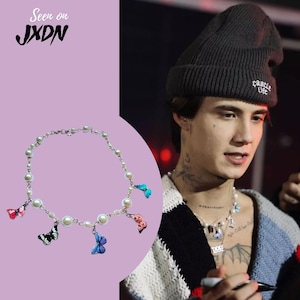 Op de afbeelding: Een zilveren ketting met witte parels en kleurrijke vlinderbedels. De ketting wordt gedragen door een persoon met een zwarte beanie en een tatoeage op hun nek.