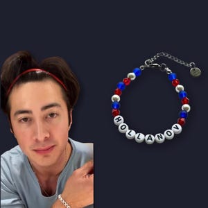 May include: A beaded bracelet with the name "HOLLANOV" spelled out in white letter beads. The bracelet features red, blue, and silver beads, and a silver chain clasp. The background is a dark navy blue.