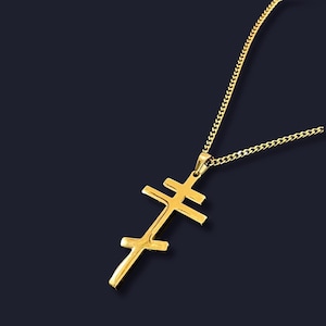 Könnte beinhalten: Eine goldfarbene Halskette mit Anhänger, die ein Doppelbalkenkreuz-Design aufweist. Das Kreuz hängt an einer passenden Goldkette. Die Halskette wird vor einem dunklen Hintergrund präsentiert, um die Details des Schmucks hervorzuheben.