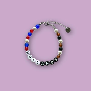 May include: A beaded bracelet with the word "HOLLANOV" spelled out in white and black letter beads. The bracelet features a mix of red, blue, white, black, and brown beads, and a silver-toned adjustable chain.