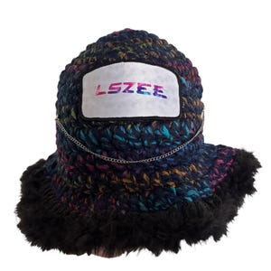 May include: A handmade, multi-colored crochet bucket hat with a black fur trim. The hat features a white patch with the word "LSZEE" in colorful lettering and a silver chain detail.