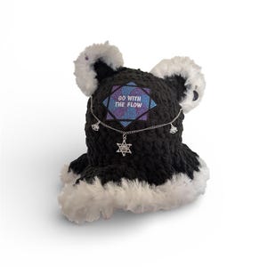 May include: Black crochet hat with white faux fur trim and ear accents. The hat features a silver chain with charms and a pendant. A patch on the front reads "GO WITH THE FLOW" in white text against a blue and purple background.