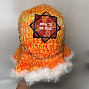 Puede incluir: Un gorro tejido a mano, naranja y amarillo, con un ribete de piel sintética blanca. El gorro presenta un parche decorativo con el texto "GO WITH THE FLOW" y una cadena plateada. Un accesorio de moda único.
