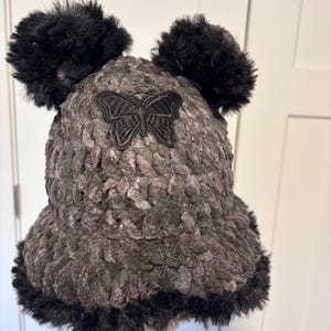 May include: A dark gray crochet bucket hat featuring a black embroidered butterfly patch on the front. The hat is topped with two fluffy black pom-poms and finished with a thick black faux fur trim along the edge of the brim.