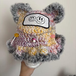 May include: A crochet bucket hat featuring a multi-colored yarn blend of yellow, pink, and white with a fuzzy grey trim. The hat includes two ear-like protrusions, a white patch with a black wavy smiley face, and a silver chain with mushroom and lollipop charms.