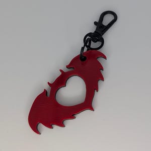 May include: A red keychain with a heart-shaped cutout and a jagged edge design. The keychain has a black metal clasp.