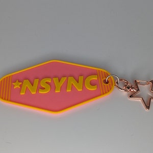 May include: A pink and yellow keychain with the word "*NSYNC" on it. The keychain is shaped like a motel sign and has a gold star charm attached.