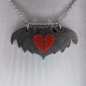 Puede incluir: Un collar con un colgante en forma de murciélago negro y un corazón roto rojo en el centro. El collar está hecho de una cadena plateada.