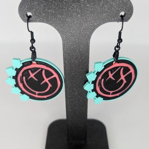 May include: A pair of dangle earrings with a black smiley face design on a turquoise background. The smiley face is outlined in pink. The earrings are hanging from a black earring hook.