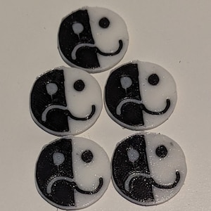 Puede incluir: Cinco fichas redondas blancas y negras con un diseño de cara dividida. Un lado de cada ficha tiene una cara triste y el otro lado tiene una cara feliz.