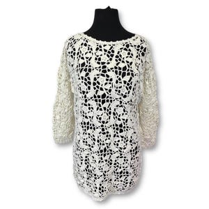 May include: Off-white crochet top with three-quarter sleeves. The openwork design features a floral pattern. The top is displayed on a black mannequin. The crochet top is a stylish addition to any wardrobe.
