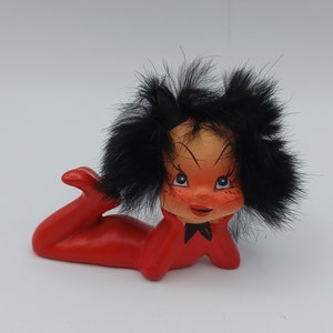 Può includere: Una statuetta di ceramica rossa di un diavolo con capelli e corna neri. La statuetta è sdraiata di lato con la testa appoggiata sulla mano.
