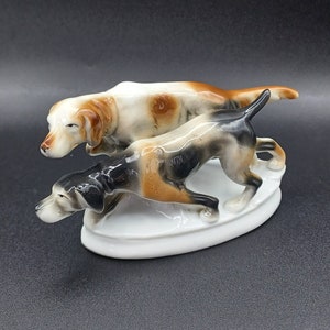 Puede incluir: Una figurita de porcelana de dos perros de caza en posición de carrera. Los perros son marrones, blancos y negros. La figurita está sobre una base ovalada blanca.