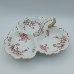 Puede incluir: Un plato de servir de porcelana blanca con tres secciones y un asa dorada. El plato está decorado con un patrón floral de rosas rosas y flores moradas.