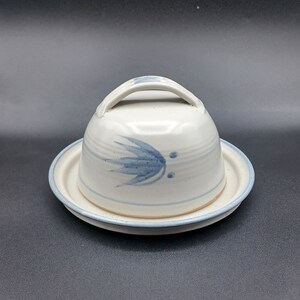 May include: A white ceramic butter dish with a blue floral design and a lid with a handle. The dish is on a white plate.