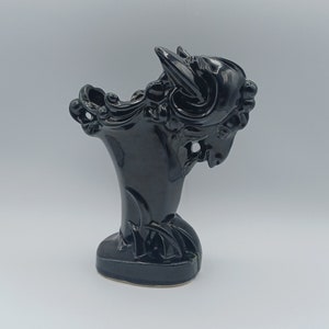 May include: A black ceramic vase shaped like a ram's head. The vase has a glossy finish and detailed features, including the ram's horns, face, and flowing mane. The base is rectangular and the design is artistic.