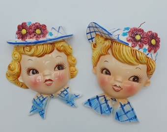 Puede incluir: Dos placas de pared de cerámica vintage que representan las caras de una niña y un niño. Ambas figuras llevan sombreros con detalles florales y ropa a cuadros azul y blanca.