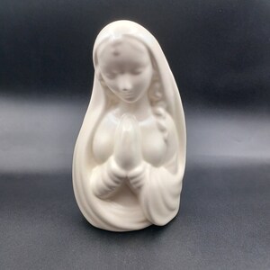 May include: White ceramic figurine of a woman in a prayer pose, wearing a long robe. The figurine is a religious figure, possibly Mary, the mother of Jesus.