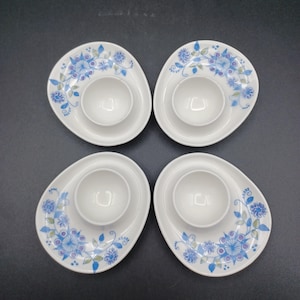 May include: A set of four white ceramic egg cups, each featuring an asymmetrical oval base decorated with a blue and green floral pattern. The central cup is circular and raised, designed to hold a single egg, with a smooth, glossy finish.