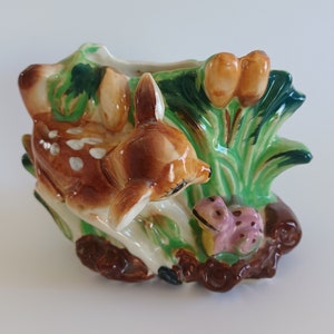 May include: A ceramic planter shaped like a deer and a frog in a woodland setting. The deer is brown with white spots and the frog is pink with brown spots. The planter is decorated with green leaves and brown branches.