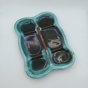 Puede incluir: Un plato de cerámica azul turquesa con cinco compartimentos. El plato tiene un borde ondulado y un palo de madera marrón descansando en dos de los compartimentos.