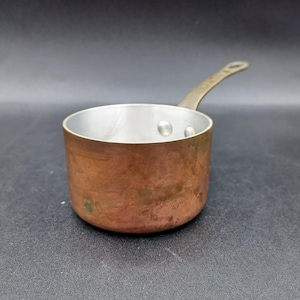May include: A small copper saucepan with a silver interior and a brass handle. The pot has a rounded base and a single, long handle. The copper exterior shows signs of age and use, with a slightly tarnished appearance. The handle is attached with two rivets.