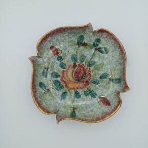 May include: A small, decorative ceramic dish with a scalloped edge. The dish is painted with a floral design in shades of green, red, and yellow. The design features a large rose with leaves and stems.