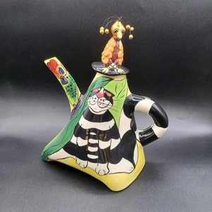 Könnte beinhalten: Eine Teekanne aus Keramik mit einem schwarz-weißen Katzendesign. Die Teekanne hat ein grünes, gelbes und orangefarbenes Blumendesign und eine kleine Vogelfigur oben drauf.