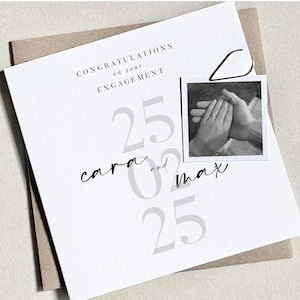K&ouml;nnte beinhalten: Eine wei&szlig;e Verlobungskarte mit dem Text "CONGRATULATIONS on your ENGAGEMENT" und den Namen "cara and max". Ein kleines Schwarz-Wei&szlig;-Foto zeigt zwei H&auml;nde mit einem Verlobungsring. Die Karte liegt auf einem braunen Umschlag.