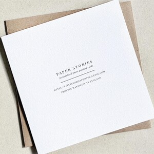 K&ouml;nnte beinhalten: Eine wei&szlig;e Gru&szlig;karte mit dem Text "PAPER STORIES personalized photo greeting cards" und der Website-Adresse. Die Karte liegt auf einem braunen Umschlag mit strukturierter Oberfl&auml;che.