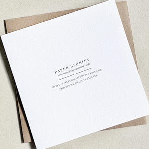 Może przedstawiać: Biała kartka z pozdrowieniami z tekstem "PAPER STORIES personalized photo greeting cards" i adresem strony internetowej. Karta leży na brązowej kopercie o teksturowanej powierzchni.