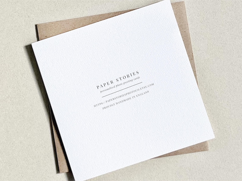 Może przedstawiać: Biała kartka z pozdrowieniami z tekstem "PAPER STORIES personalized photo greeting cards" i adresem strony internetowej. Karta leży na brązowej kopercie o teksturowanej powierzchni.