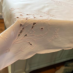 以下が含まれることがあります： 淡いピンク色のテーブルクロスで、縁に複雑な白い花柄の刺繍とカットワークのディテールが施されています。生地は滑らかな質感で、繊細に縫い込まれた花模様と、角から垂れ下がるスカラップ状の縁取りが特徴です。