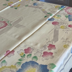 May include: A cream-coloured tablecloth featuring a repeating pattern of large pink, blue, and yellow flowers with green leaves. The fabric includes light grey geometric shapes and lines that intersect with the floral motifs across the surface.