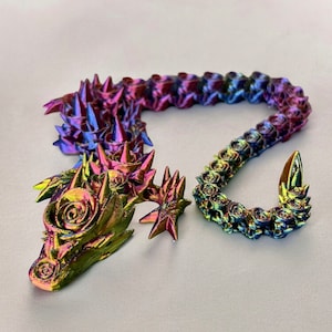 May include: A 3D printed iridescent dragon sculpture with a rose-like design. The dragon is curled up with its tail wrapped around its body. The sculpture is made of a shiny, metallic material.