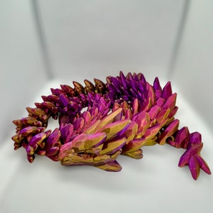 May include: A 3D printed dragon sculpture made of purple and gold plastic. The dragon is curled up in a sleeping position.