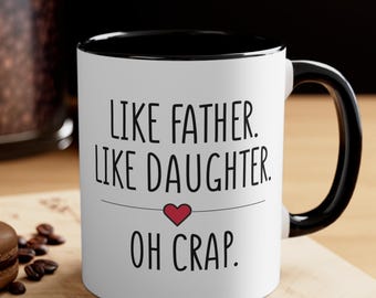 May include: White ceramic mug with a black interior and handle. The mug features the text "LIKE FATHER. LIKE DAUGHTER. OH CRAP." in black lettering, with a small red heart. The mug is on a wooden surface.