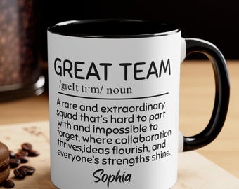 May include: A white ceramic mug with a black interior and handle. The mug features the words "GREAT TEAM" in large black letters, along with a definition and the name "Sophia" at the bottom. The mug is on a wooden surface.
