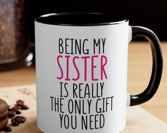 May include: White ceramic mug with a black interior and handle. The mug features the text "BEING MY SISTER IS REALLY THE ONLY GIFT YOU NEED" in black and pink lettering. The mug is on a wooden surface.