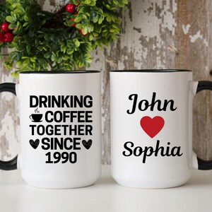May include: Two white coffee mugs with black handles and black rims. One mug says "Drinking Coffee Together Since 1990" with a coffee cup icon and two hearts. The other mug says "John" with a red heart and "Sophia".